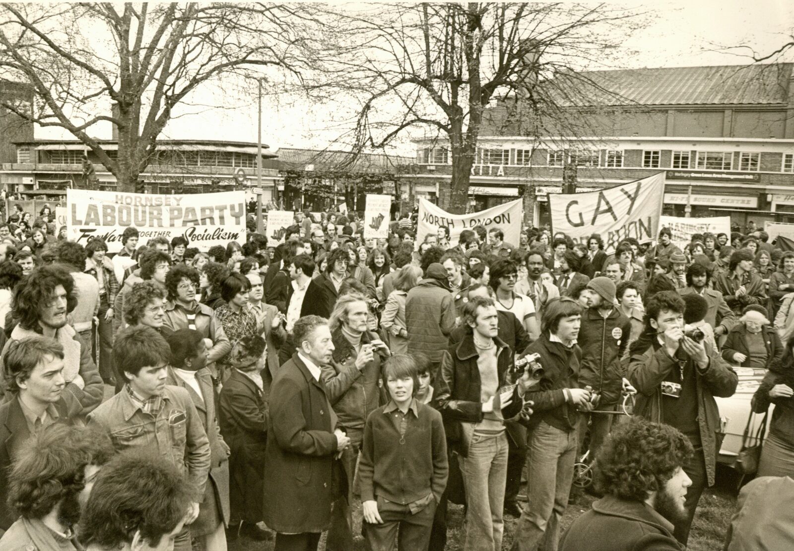 A large crowd gathered under bare trees at the 1977 Wood Green demonstration, with Hornsey Labour Party and gay liberation banners among the placards.